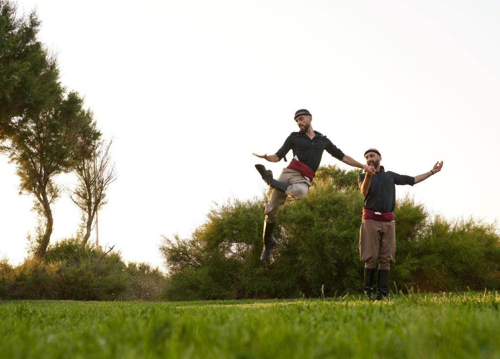 Divi dejotāji izpilda tradicionālo deju laukā, viens no viņiem veic gaisa lēcienu. Fons ir zaļš zālājs ar kokiem un krūmiem.