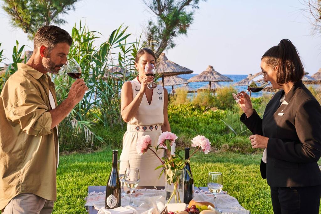 Vīna degustācija ārā pie jūras, kur trīs cilvēki, tērpti eleganti, izbauda sarkanvīnu un priecājās par dabu. Uz galda atrodas vīna pudeles un ziedi.