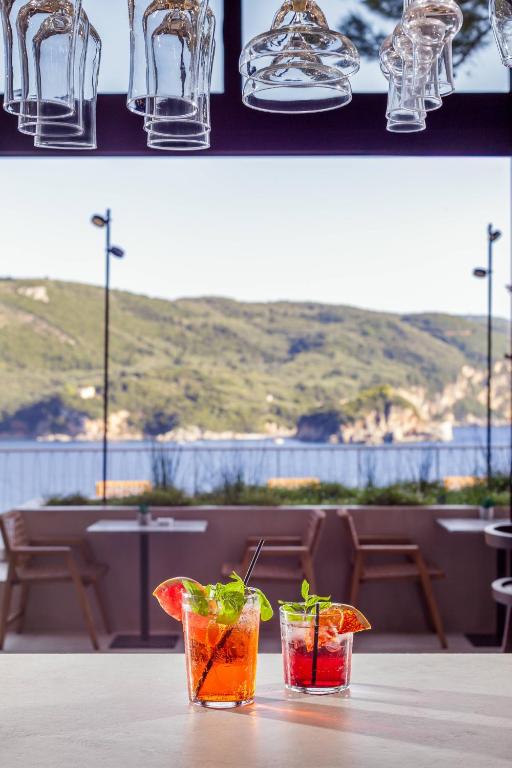 Divi kokteiļi uz galda ar ainavu, kas ietver kalnus un jūru. Aiz galda redzami koka krēsli un lieli logi, caur kuriem ieplūst dabiska gaisma.