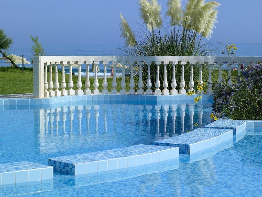 Baseins ar skaidru ūdeni un dekoratīvu balustrādi, apkārtne ar zaļām zālēm un ziediem. Jūras fons un saulaina debess rada mierīgu atmosfēru.
