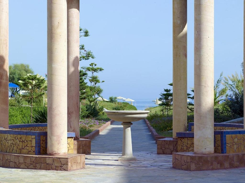 Aizsargājoša arhitektūra ar kolonnām un ainavu, kurā redzams dārzs un jūra. Šī ainava rada mierīgu un relaksējošu atmosfēru.