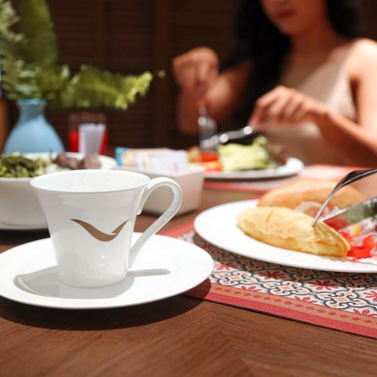 Balta kafijas krūze ar stilizētu zīmi uz šķīvja, blakus ir dažādi ēdieni, tostarp omlete un salāti, radot patīkamu brokastu atmosfēru.