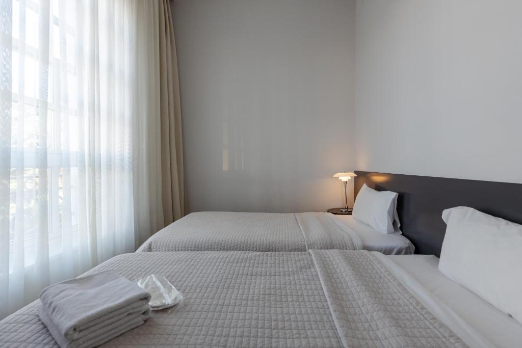 Gaisma un mūsdienīgs guļamistabas interjers ar divām atsevišķām gultām, baltiem gultas piederumiem un logiem ar caurspīdīgām aizkariem. Uz galda redzama nakts lampiņa.