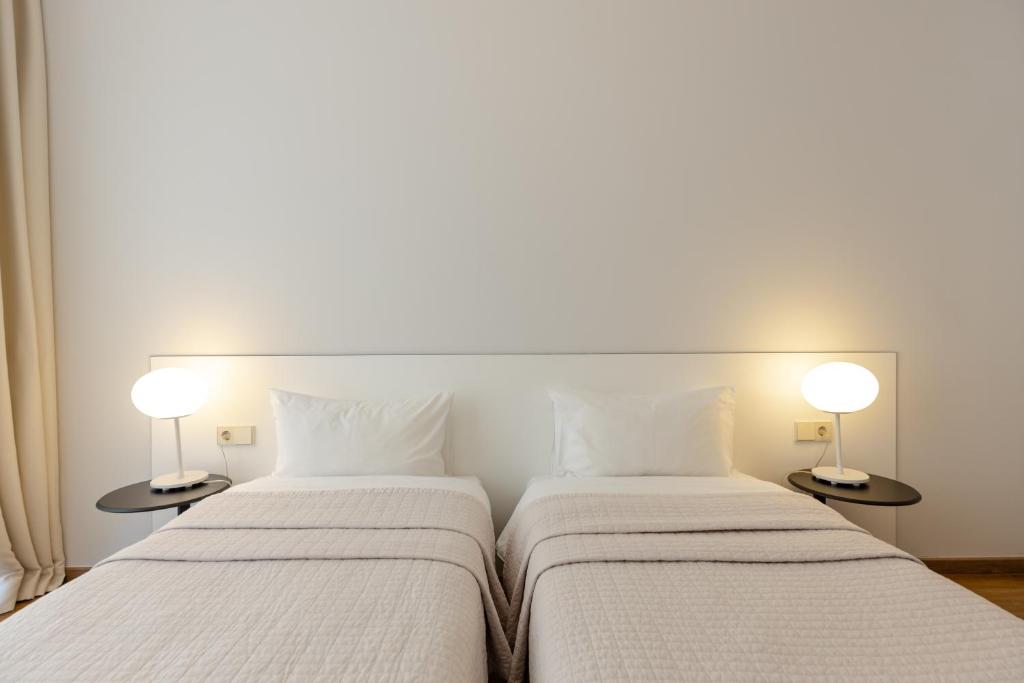 Gaiša guļamistaba ar divām atsevišķām gultām, mīkstiem spilveniem un modernām lampām blakus katrai gultai. Sienas ir neitrālas krāsas, radot mierīgu atmosfēru.