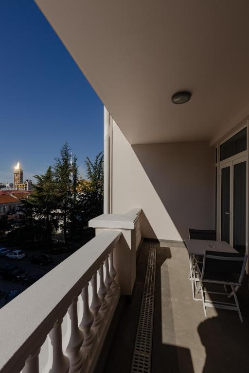 Balkons ar skatu uz pilsētu, kurā redzama torņa gaisma un skaidrs zilas debesis. Balta balustrāde un vienkārša mēbele piešķir mūsdienīgu atmosfēru.