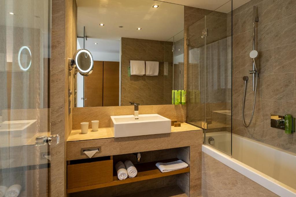 Mūsdienīgs vannas istabas interjers ar stikla dušu un elegantu izlietni, apvienojot silta akmens toņus un modernu apgaismojumu. Iekļauti arī dvieļi un higiēnas līdzekļi.