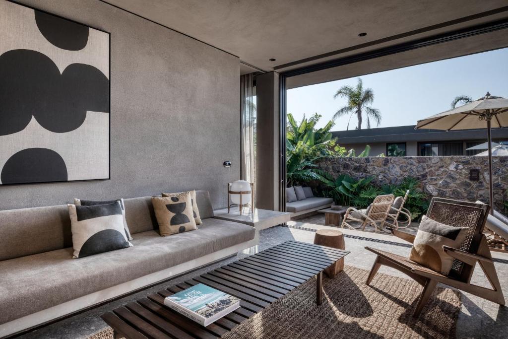 Mūsdienīgs viesistabas interjers ar pelēku dīvānu, melnbaltu mākslas darbu un atvērtu skatu uz terasi, kas ierāmēta ar zaļām palmām un augiem. Tajā ir koka galdiņš un dažādi dekoratīvie spilveni.