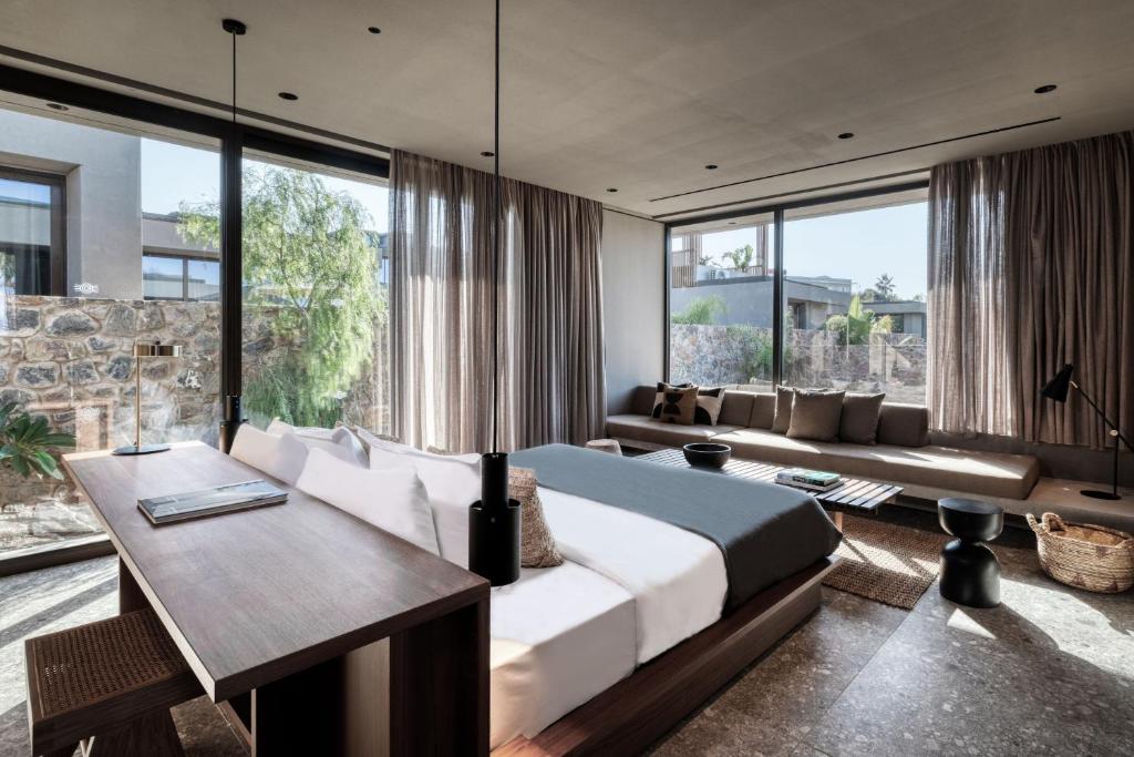 Mūsdienīga guļamistaba ar lieliem logiem, dabiskiem elementiem un minimālistisku dizainu, kas rada mierīgu un relaksējošu atmosfēru. Interjerā redzama ērta gulta, dīvāns un koka galds.