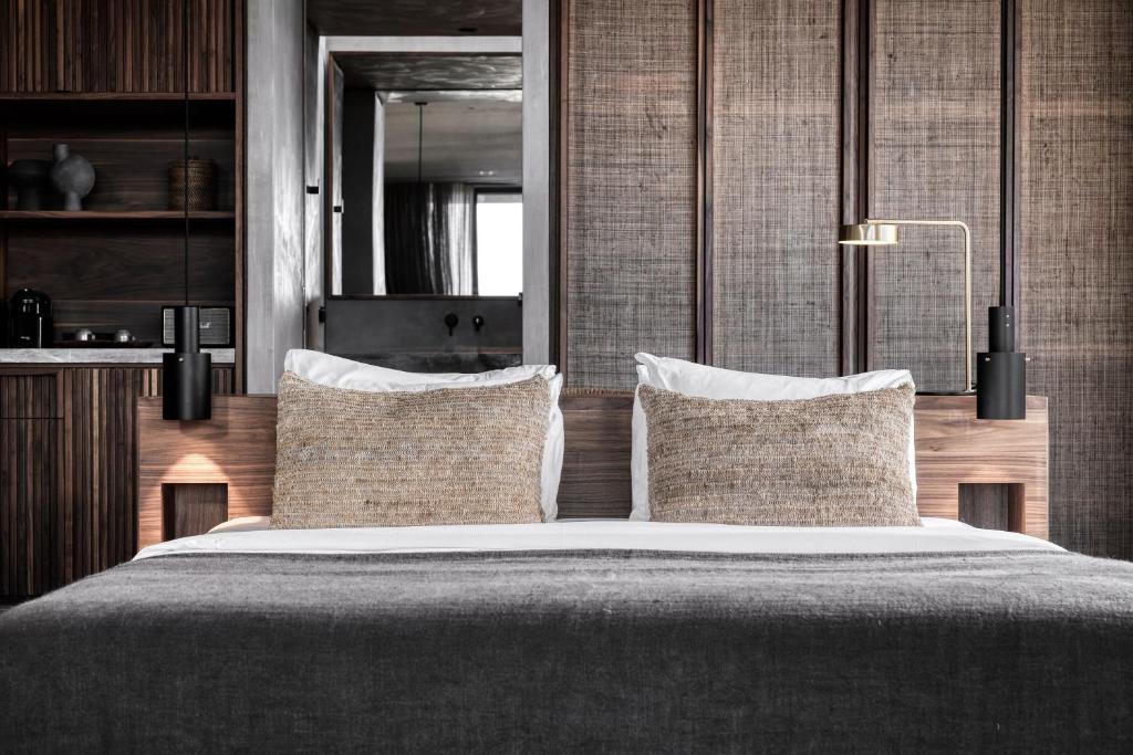 Mūsdienīgs guļamistabas interjers ar divām dekoratīvām spilvenēm uz gultas un elegantu apgaismojumu. Sienas klātas ar teksturētu materiālu, radot siltu un aicinošu atmosfēru.