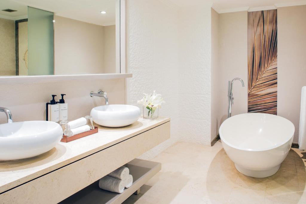 Mūsdienīgs vannas istabas interjers ar divām izlietnēm, brīvo stāvošu vannu un dekoratīvu augu. Eleganta dizaina elementi un neitrālas krāsas rada relaksējošu atmosfēru.