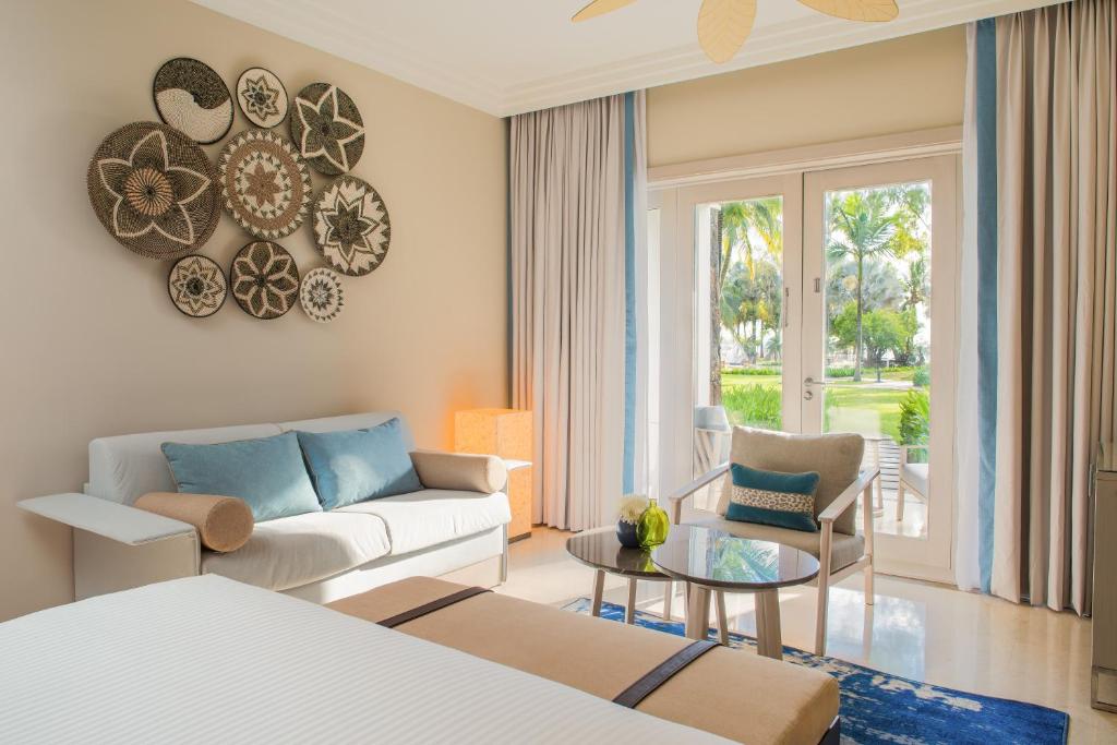 Mūsdienīgs viesnīcas numurs ar stilīgu interjeru, kurā ir ērta sofa, krēsls un liels logs, kas ved uz dārzu. Uz sienas ir dekoratīvi grozi, kas piešķir telpai unikālu raksturu.