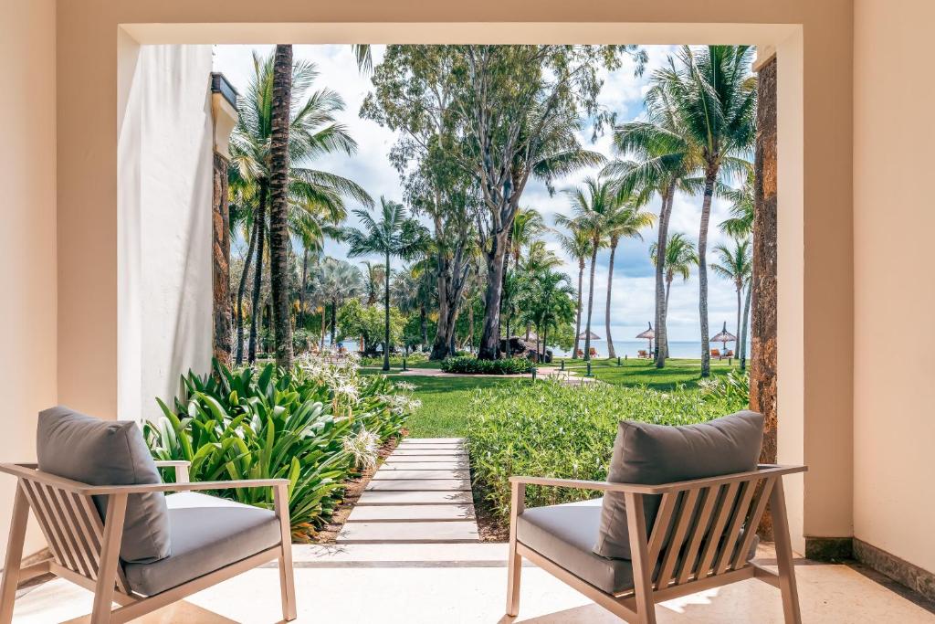 Skats no terases uz skaistu dārzu ar palmām un ziediem, kas ved uz pludmali. Divas ērtas krēsli ir novietoti blakus, radot mierīgu un relaksējošu atmosfēru.