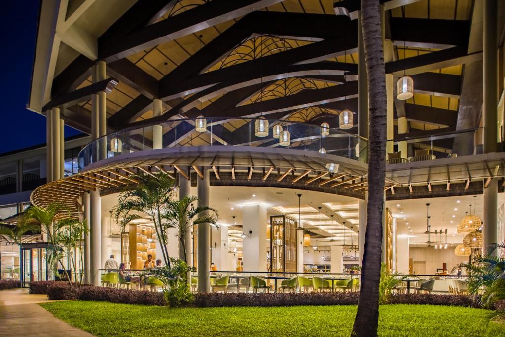 Moderns restorāns ar plašām terasēm un elegantiem gaismas ķermeņiem, kas apgaismo telpu naktī. Apkārtne ir zaļa ar palmām un zālāju, radot relaksējošu atmosfēru.