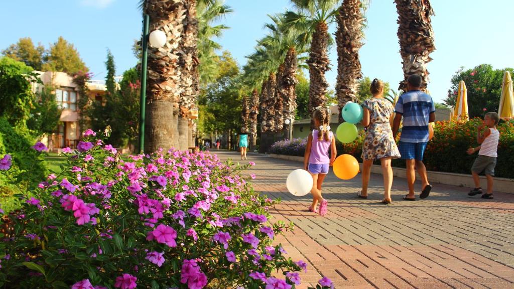 Kāds saulains parks ar ziediem un palmām, kur ģimene pastaigājas, turēdama krāsainus balonus. Bērni iepriecina ar spēlēšanos un dzīvesprieku.