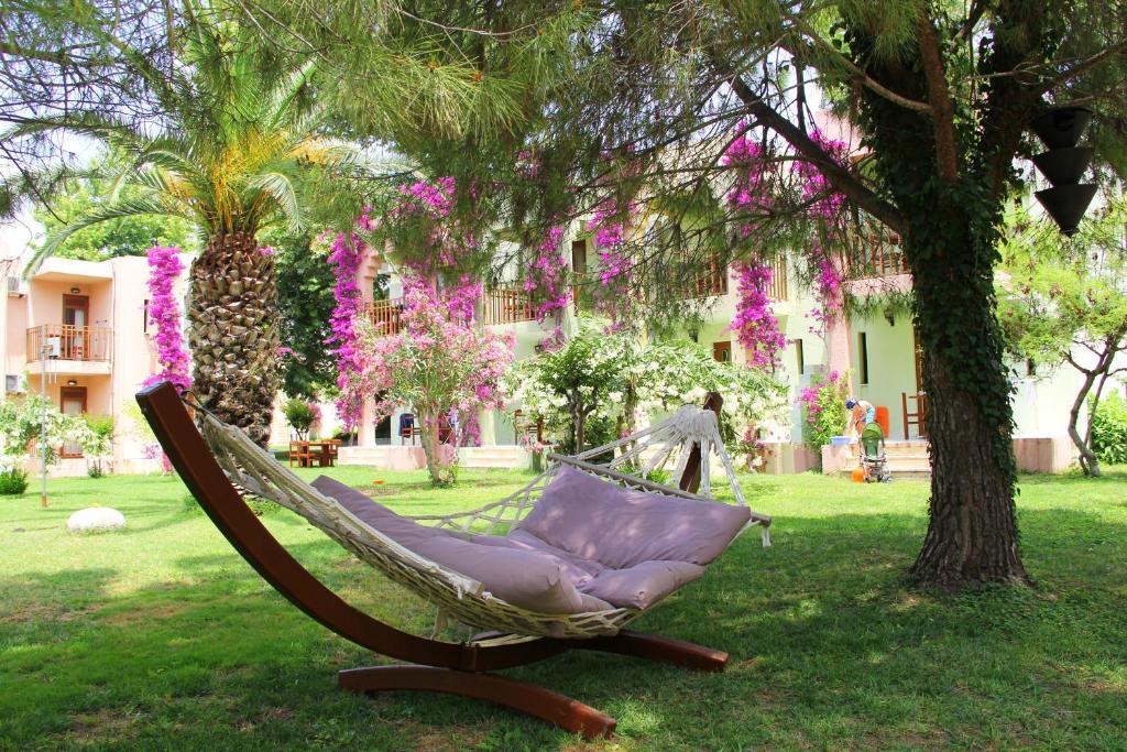 Mierīga dārza aina ar šūpuļtīklu, kas novietots zālienā, ko ieskauj ziedoši augi un zaļumi. Fons veido mūsdienīga ēka ar balkoniem, radot relaksējošu atmosfēru.