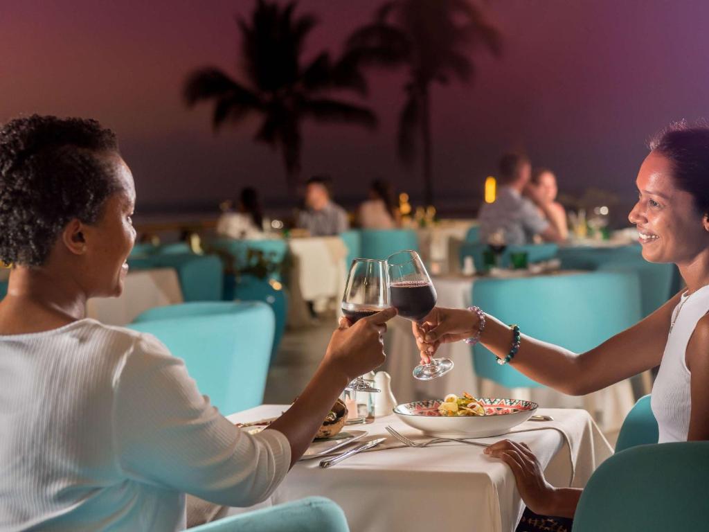 Divas sievietes, sēžot restorānā ar skatu uz saulrietu, uzmundrina, dzerot sarkanvīnu un smejoties. Apkārt ir eleganta atmosfēra ar baltiem galdiem un ziliem krēsliem.