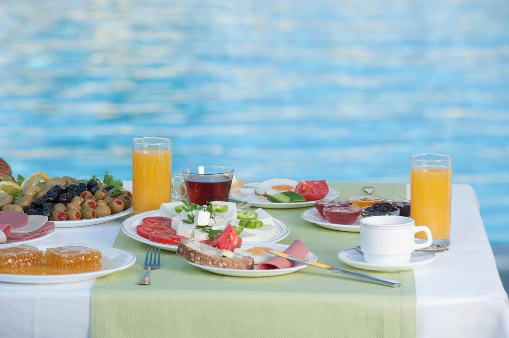 Bagātīgs brokastu galds ar dažādiem ēdieniem, tostarp olīvām, tomātiem, maizi un dzērieniem, dekorēts pie ūdens.