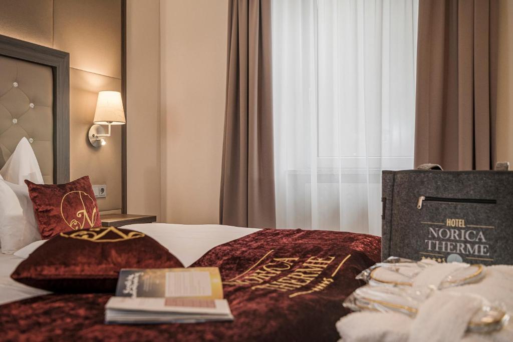 Mūsdienīgs viesnīcas numurs ar elegantu interjeru, kas ietver sarkanas krāsas gultas veļu un spilvenus. Blakus ir viesnīcas soma un brošūra, kas piedāvā informāciju par viesnīcu Norica Therme.