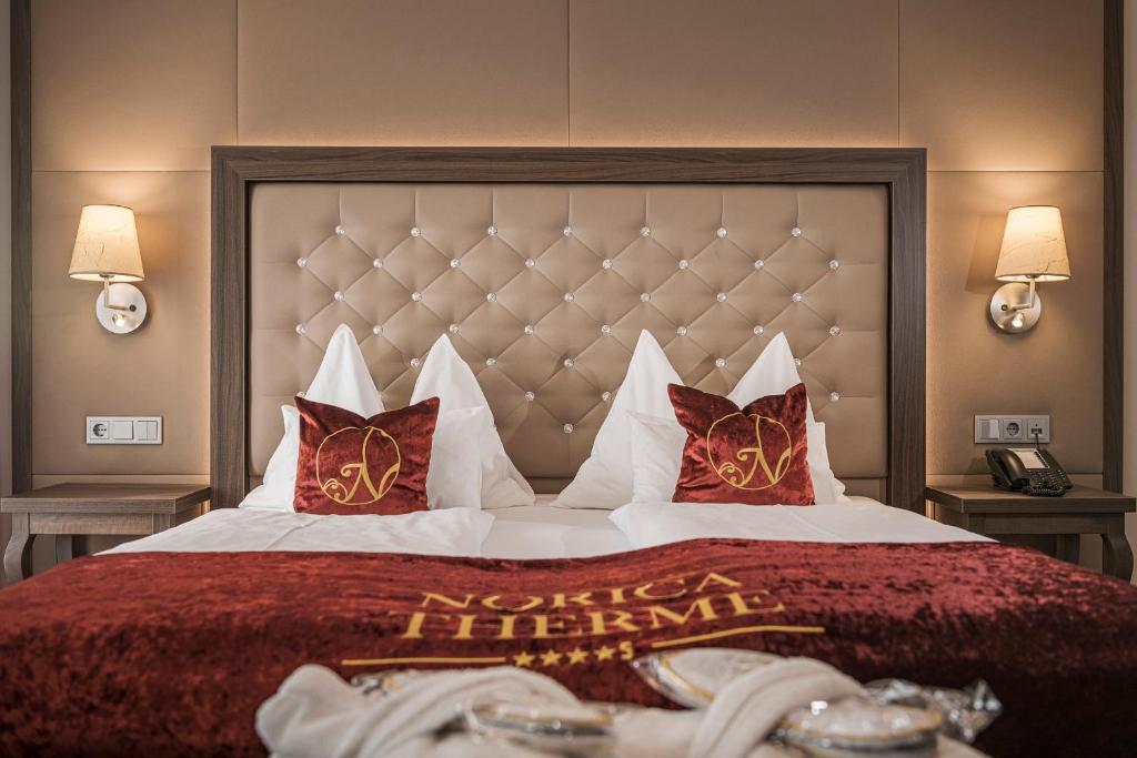 Elegants viesnīcas numurs ar gultu, dekorētu ar sarkanu samtu un spilveniem ar viesnīcas logotipu. Telpā ir mūsdienīgi gaismekļi un koka mēbeles, radot komfortablu atmosfēru.