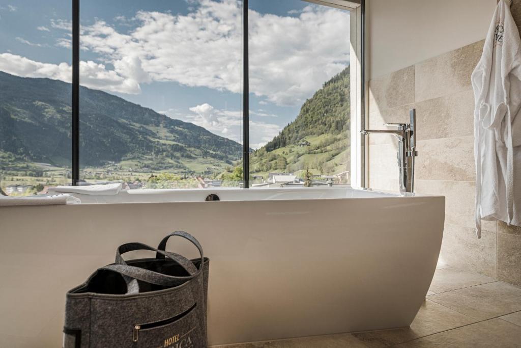 Mūsdienīga vannas istaba ar panorāmas skatu uz kalniem, baltu vannu un dabīgu apgaismojumu. Blakus ir melna soma un baltas dvieļi.