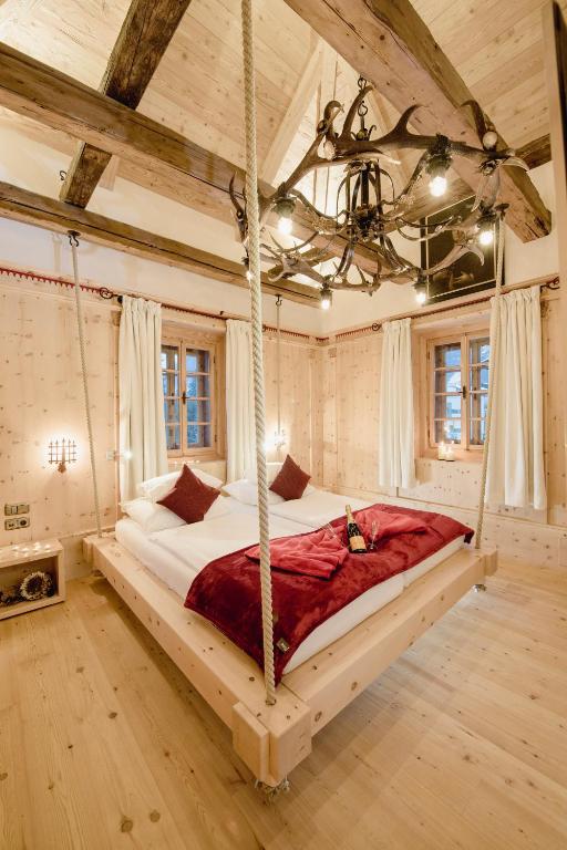 Mūsdienīgs guļamistabas interjers ar koka sijām, šūpojošu gultu un siltu apgaismojumu, radot mājīgu un romantisku atmosfēru. Iekļauti logi ar aizkariem un eleganta gultas veļa.