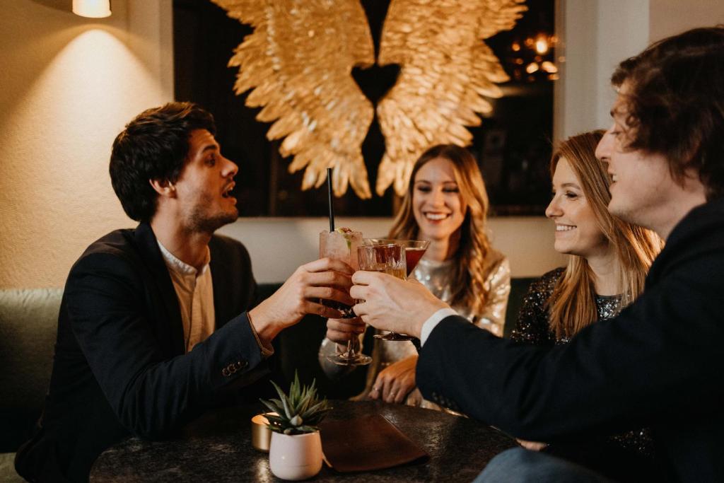 Četri cilvēki svinīgi pacelt glāzes, baudot dzērienus stilīgā bārā ar mūsdienīgu interjeru. Fons rotāts ar zeltainām spārnu formām, radot svinīgu atmosfēru.