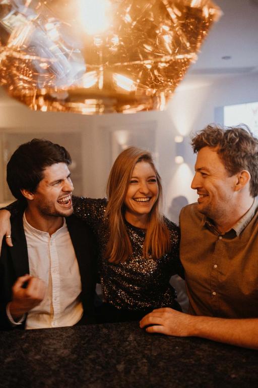 Trīs cilvēki sēž pie galda, priecīgi sarunājoties un smejoties. Viņi ir ģērbušies svinīgi, un fons ir elegants ar modernu apgaismojumu.