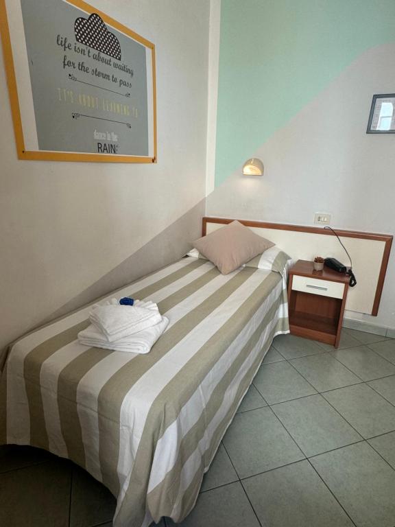 Vienkārša viesnīcas istaba ar gultu, kas pārklāta ar svītrainu gultasveļu, un piegulošu naktsgaldiņu. Uz sienas redzams iedvesmojošs citāts, kas aicina izbaudīt dzīvi.