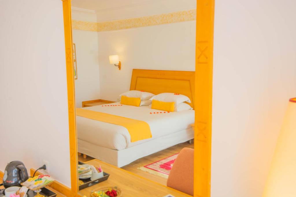 Eleganta viesnīcas numura interjers ar dzeltenu gultas pārklāju un mīkstiem spilveniem, atspoguļots lielā spogulī. Numurā ir arī gaismas un koka grīda, radot siltu un mājīgu atmosfēru.