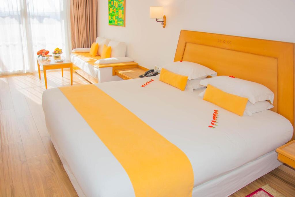 Mūsdienīga viesnīcas istaba ar saulainu interjeru, kurā ir liels gultas rāmji ar spilgti dzelteniem akcentiem un ērta sēžamā zona. Gaisma plūst cauri logiem, radot patīkamu atmosfēru.