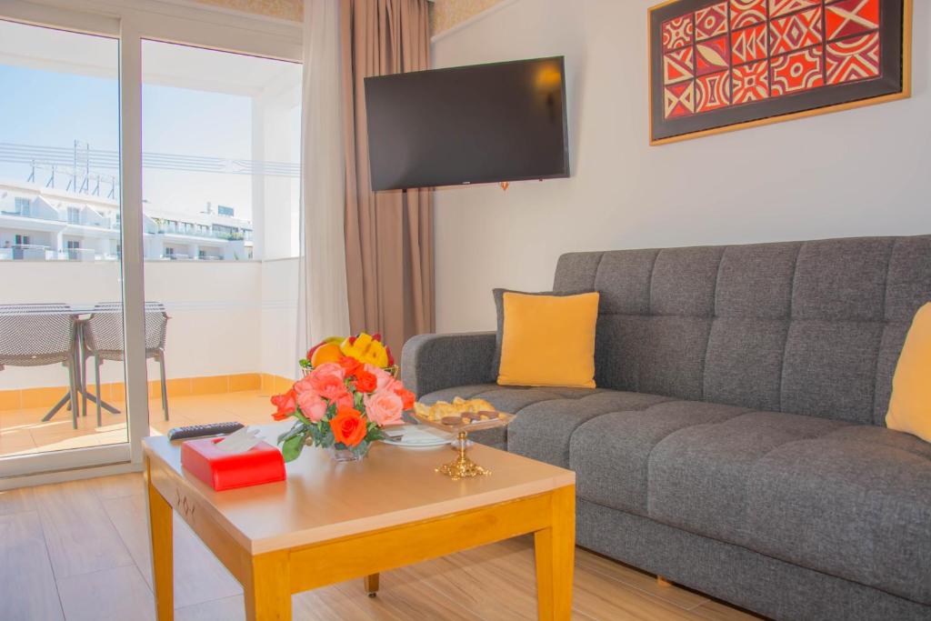 Mūsdienīgs viesnīcas numurs ar pelēku dīvānu, koka galdu un izsmalcinātu ziedu kompozīciju. Logi sniedz gaismu un skatu uz ārpusi.