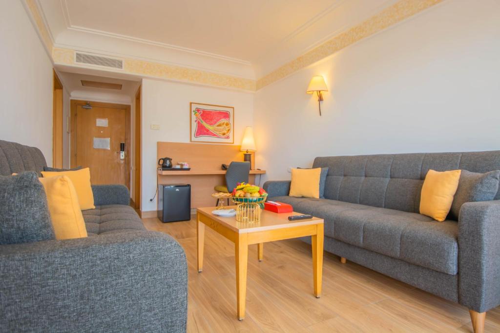 Mūsdienīga viesnīcas viesistaba ar pelēkiem dīvāniem un koka galdu, uz kura atrodas augļi un dzērienu trauki. Sienās ir gaismas un mākslas darbi, radot patīkamu atmosfēru.