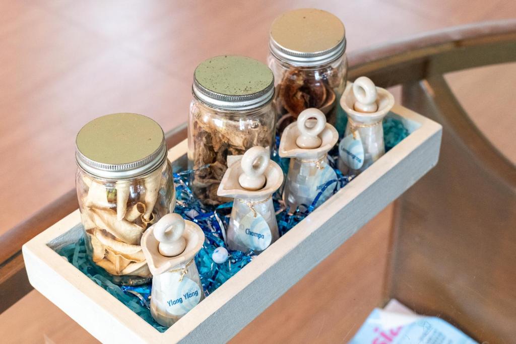 Stikla burkas ar dabīgām sastāvdaļām, piemēram, zālēm un garšvielām, izvietotas uz dekoratīvas koka paletes ar zilu papīra pildījumu.