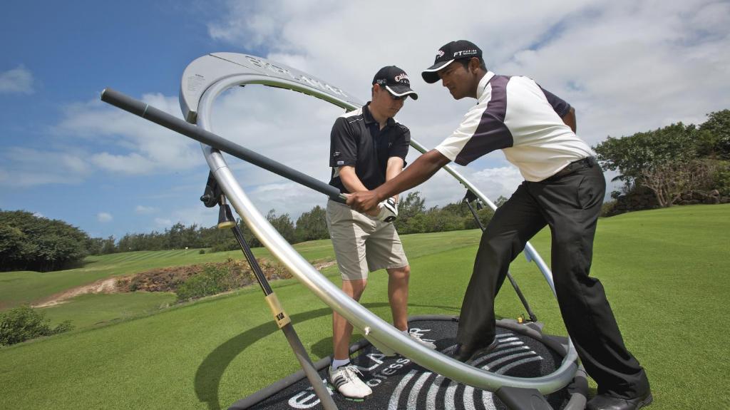 Divi golfa spēlētāji, viens no viņiem saņemot instrukcijas, izmanto speciālu treniņu aprīkojumu uz golfa laukuma. Skats uz zaļojošu ainavu un skaidru debesu fonu.