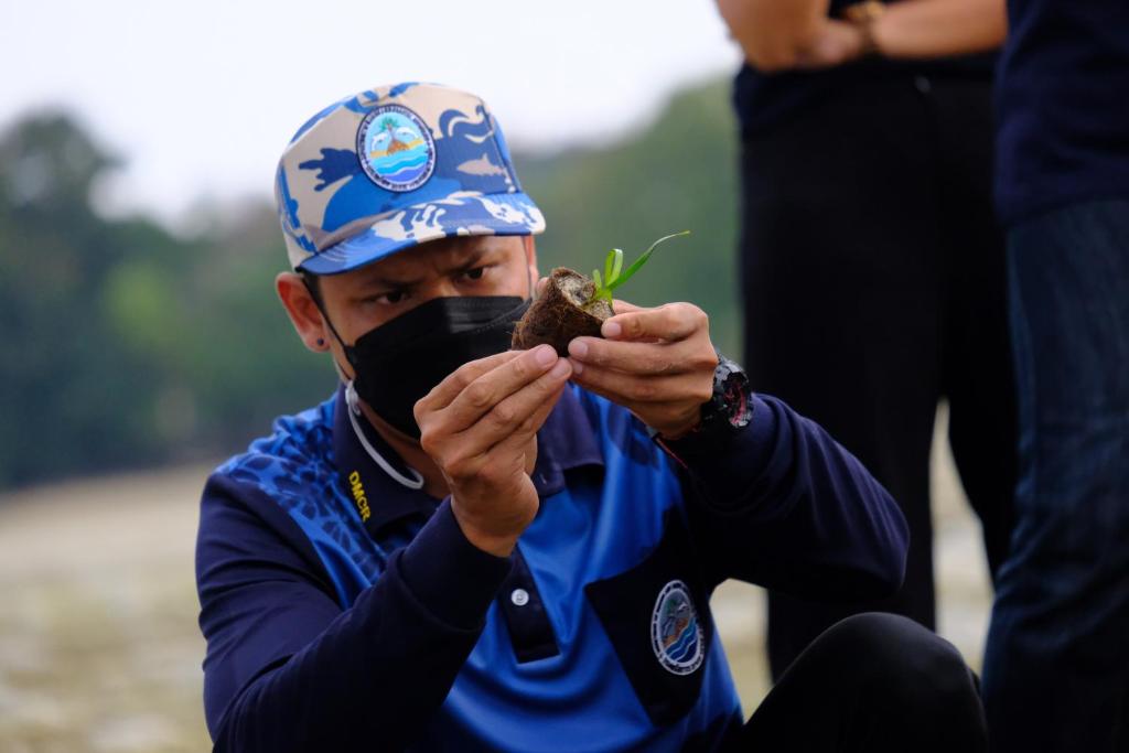 Vīrietis, tērpies zils krekls ar emblēmu un cepuri, uzmanīgi tur augu, kas izaug no zemes. Fons ir dabisks, ar cilvēkiem, kas stāv tuvumā.