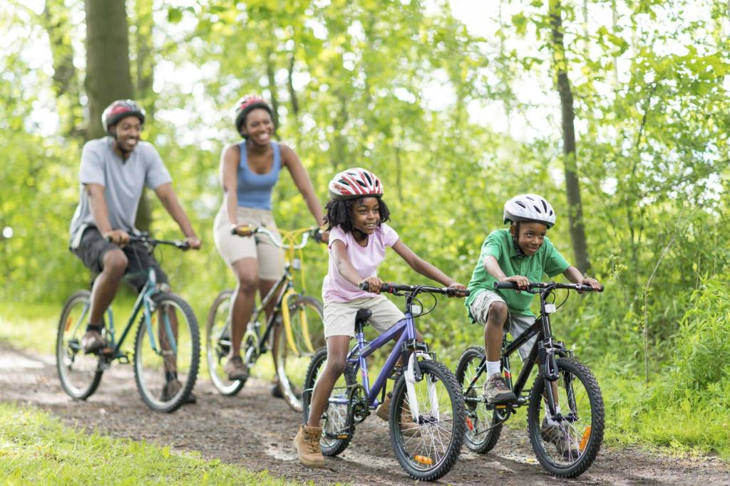 Ģimene brauc ar velosipēdiem pa zaļojošu meža taku, baudot aktīvu brīvo laiku un dabu. Bērni priecīgi brauc, valkājot ķiveres, kopā ar pieaugušajiem.