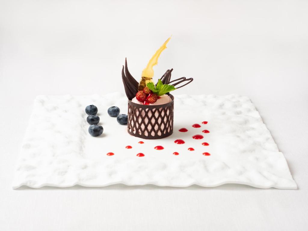 Elegants deserts ar šokolādes dekorācijām, ogām un svaigiem augļiem, pasniegts uz baltas plāksnes.