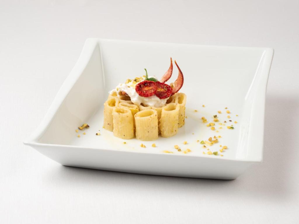 Radoša makaronu pasniegšana ar krēmīgu pildījumu, saulē kaltētiem tomātiem un riekstu sprinkles uz baltā šķīvja.