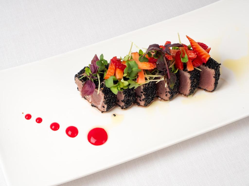 Mūsdienīga ēdiena kompozīcija ar melnajām sezama sēklām apviļātu gaļu, dekorēta ar svaigiem dārzeņiem un zemenēm, pasniegta uz baltas šķīvja ar sarkaniem mērces pilieniem.