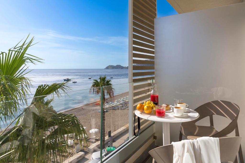 Balconā novietots galds ar brokastu komplektu, kas ietver augļus un dzērienus, skats uz pludmali un jūru ar laivām. Zāles un palmas piešķir tropisku atmosfēru.