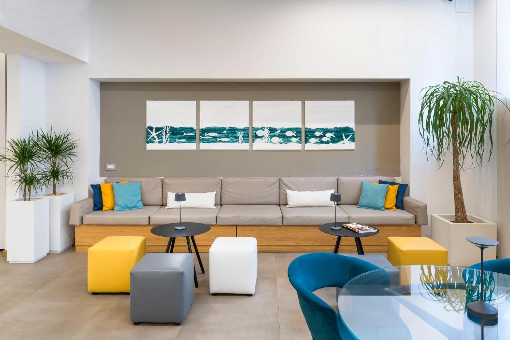 Mūsdienīgs viesnīcas interjers ar pelēku dīvānu, krāsainām spilveniem un mākslas darbiem uz sienas. Telpā ir dažādi galdi un augi, radot patīkamu un relaksējošu atmosfēru.