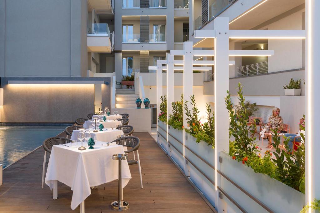 Elegants restorāna terases skats ar galdiem blakus peldbaseinam, apzaļumojums un mūsdienīga arhitektūra. Gaismas elementi rada patīkamu atmosfēru vakara baudīšanai.