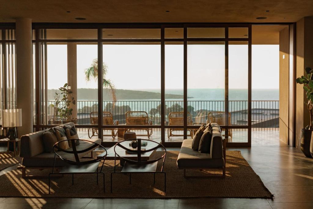 Mūsdienīgs viesistabas interjers ar plašiem logiem, kas piedāvā skatu uz jūru. Telpā ir ērti dīvāni, galds un dekoratīvie augi, radot relaksējošu atmosfēru.