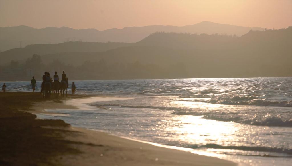 Pludmales aina ar cilvēkiem, kas pastaigājas un jājā, saulrieta gaismā, kas atspoguļojas ūdenī.