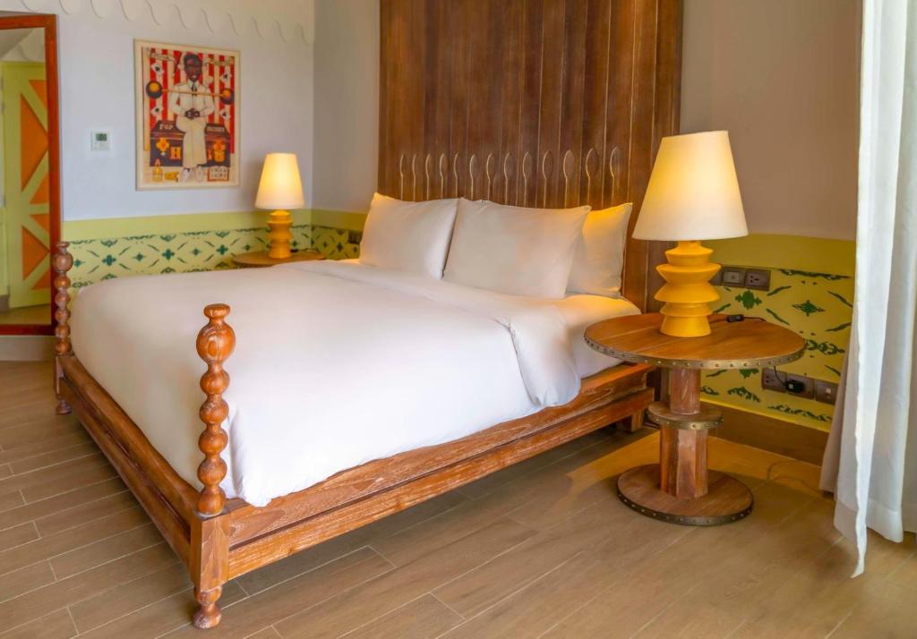 Mūsdienīgs viesnīcas numurs ar elegantu gultu, koka galdiņu un iedegtu lampu, kas rada mājīgu atmosfēru. Sienas rotā krāsains mākslas darbs, piešķirot telpai raksturu.