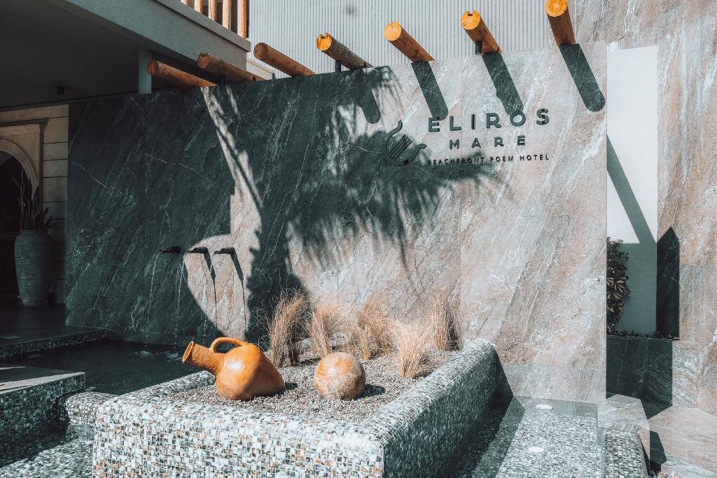 Eliros Mare viesnīcas ieeja ar modernu dizainu, akmens sienu un dekoratīvām podiņām. Redzami divi māla trauki un zaļumi, kas papildina atmosfēru.