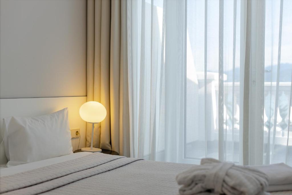 Mūsdienīgi iekārtots guļamistabas stūris ar gultu, siltu apgaismojumu un caurspīdīgām aizkariem, kas ļauj gaismai ieplūst telpā.