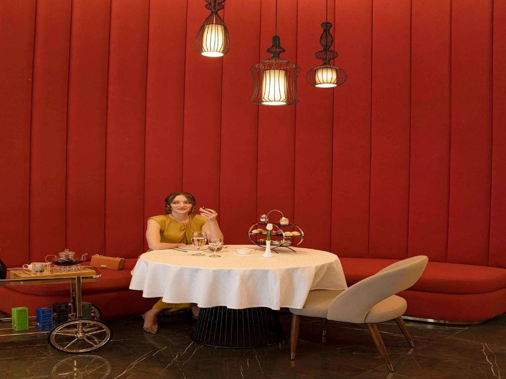 Eleganta kafejnīca ar sarkaniem sienām, kur sieviete sēž pie galda ar desertiem un dzērieniem. Gaismas spuldzes radījušas siltu atmosfēru.