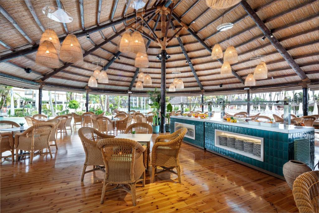 Moderns restorāns ar apaļu jumtu, koka grīdu un rattan mēbelēm, kurā redzami gaismas ķermeņi un zaļi augi. Telpa ir gaiša un eleganta, radot patīkamu atmosfēru.