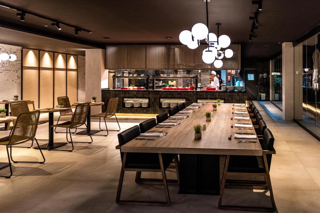 Mūsdienīgs restorāns ar elegantu interjeru, garu galdu un atvērtu virtuvi, kurā redzams šefpavārs, kas gatavo ēdienus. Gaismas elementi piešķir telpai siltu un aicinošu atmosfēru.